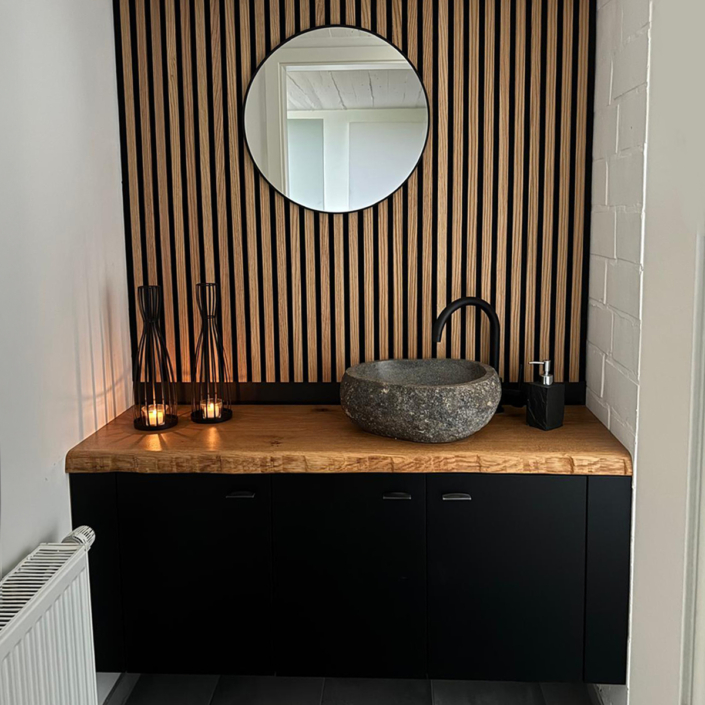 Badezimmer mit schwarzem Unterschrank, Holzarbeitsplatte, rundem Steinwaschbecken, schwarzem Wasserhahn und rundem Spiegel vor Wand mit vertikalen Holzpaneelen.